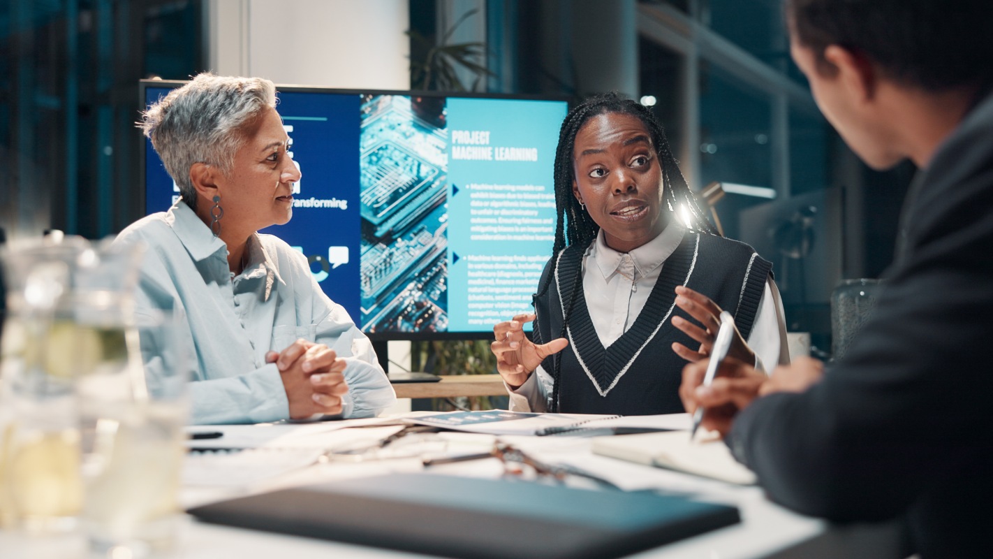 A group of professionals engages in a discussion in a modern office, with a digital screen displaying project details in the background.