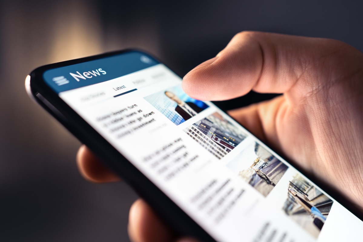 A hand holding a smartphone displaying a news app with articles and images. The screen shows headlines under the "Latest" tab, suggesting engagement with current events.
