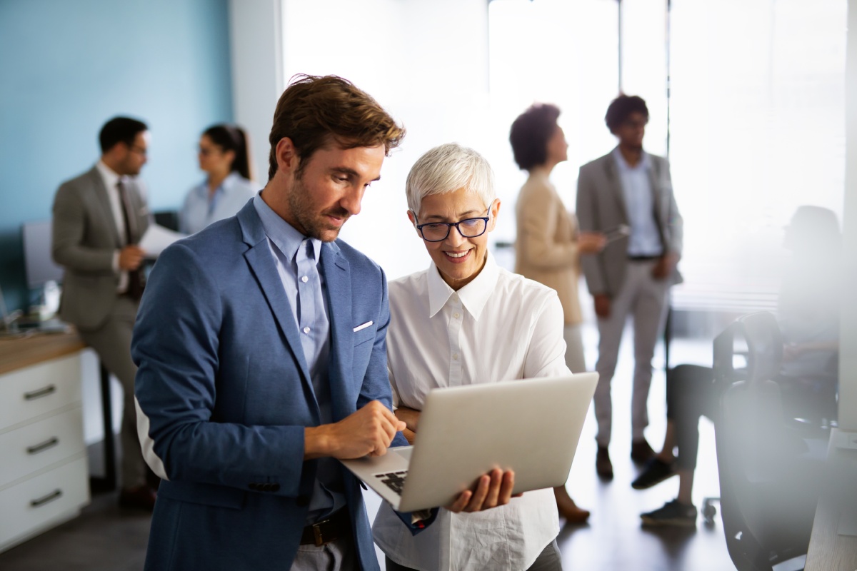 Smiling colleagues in business attire discuss work on a laptop in a sunlit office. Four others chat in the blurred background, creating a collaborative vibe.